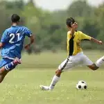Giocatore di calcio in azione durante una partita, con sullo sfondo bandiere di squadre diverse